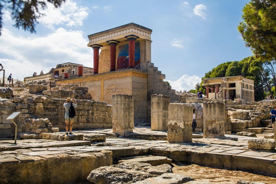 Una persona con sombrero fotografía las antiguas ruinas de piedra de un edificio con columnas rojas prominentes bajo un cielo soleado.
