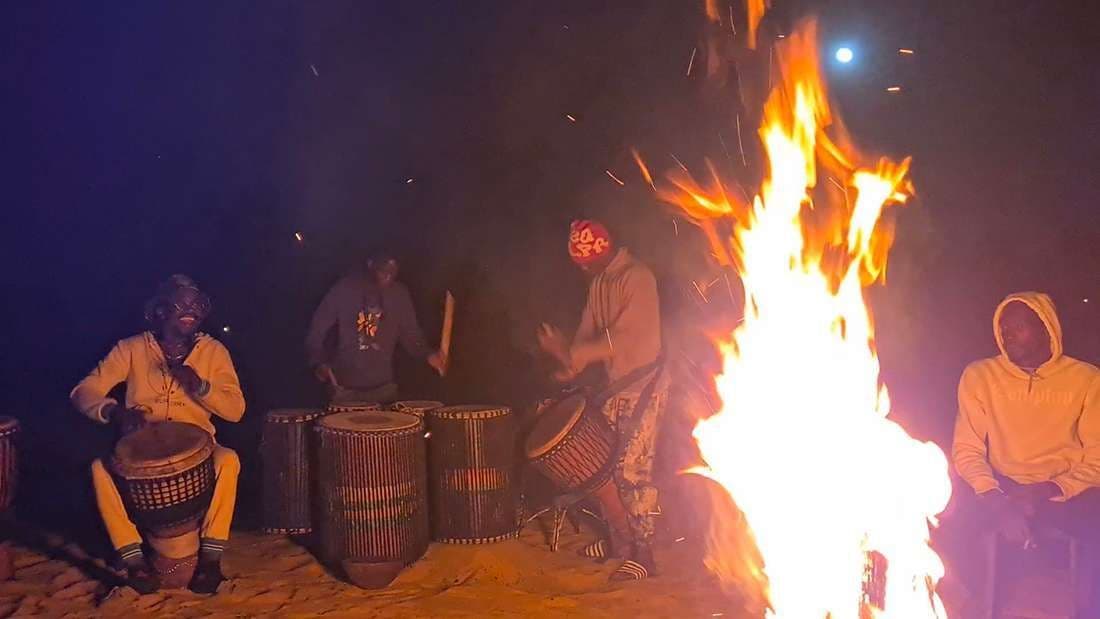 Eine WeRoad Gruppenreise: Menschen spielen nachts am großen Lagerfeuer Trommel und sitzen zusammen.