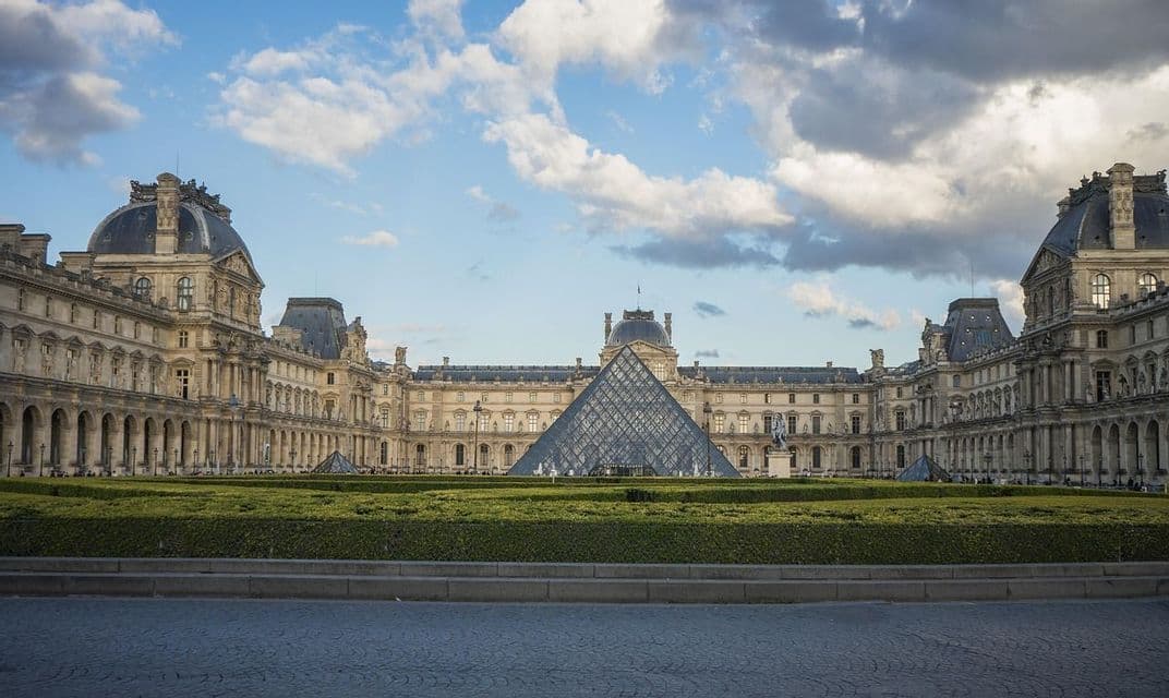 Un grand palais orné avec une pyramide de verre dans sa cour centrale, derrière une pelouse bien entretenue sous un ciel partiellement nuageux.
