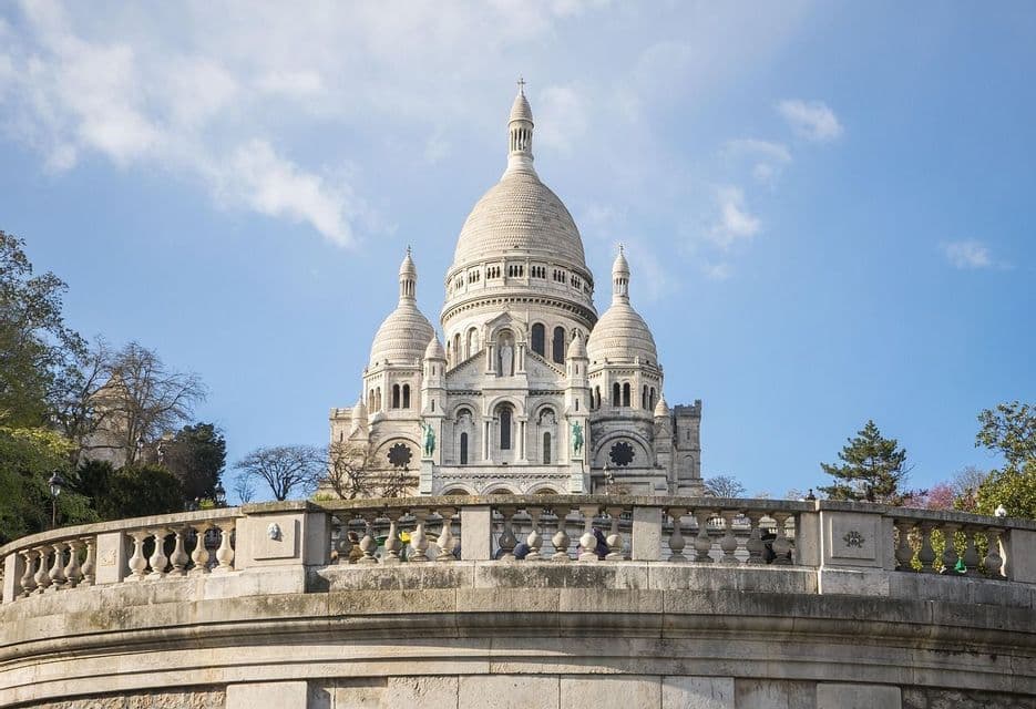 Une grande basilique blanche avec de multiples dômes se dresse derrière une balustrade en pierre sous un ciel bleu parsemé de légers nuages.