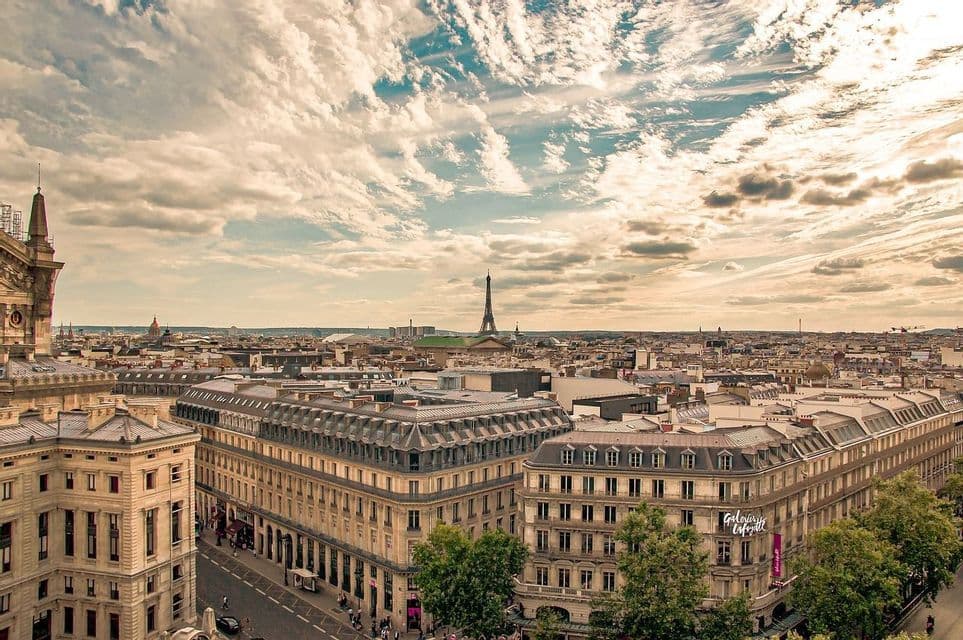Vue panoramique sur une ville, avec ses bâtiments historiques et ses toits, et une haute tour de fer se dressant au loin sous un vaste ciel nuageux.