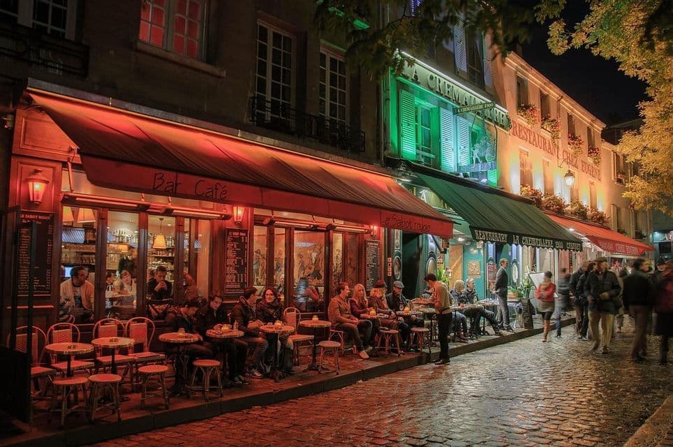 Des gens dînent en terrasse sous le chaud auvent rouge d'un café-bar, dans une rue pavée, la nuit.