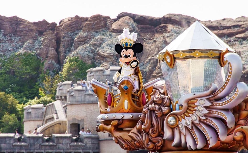 Mickey Mouse, vêtu d'un costume royal blanc et or avec une couronne, salue depuis un char de parade orné, devant des montagnes rocheuses et un château.