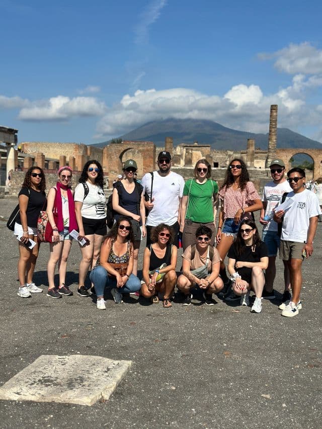 Un groupe WeRoad en voyage pose pour une photo sur une place pavée avec des ruines antiques et une grande montagne en arrière-plan.