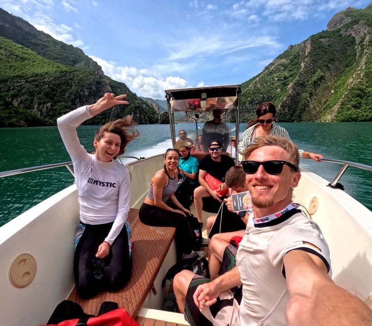 Un groupe WeRoad en voyage souriant pour un selfie sur un bateau, naviguant sur un lac entre des montagnes vertes et escarpées.