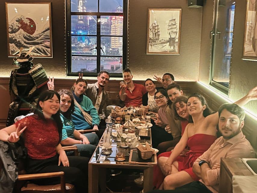 Un groupe WeRoad pose et sourit autour d'une longue table dans un restaurant, avec la ligne d'horizon de la ville visible la nuit par une fenêtre.