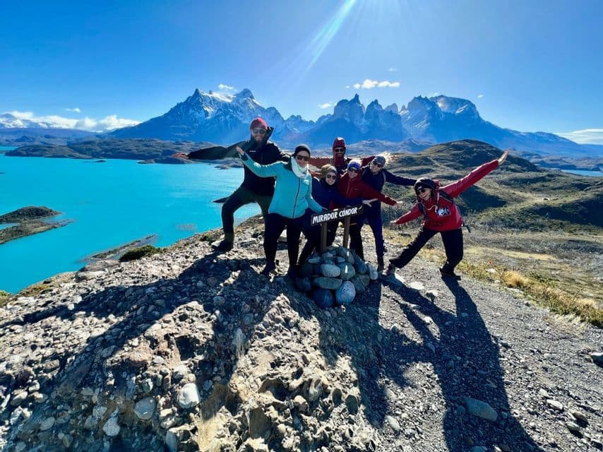 Un groupe WeRoad en voyage pose pour une photo depuis un point de vue surplombant un lac turquoise éclatant et des montagnes déchiquetées et enneigées.