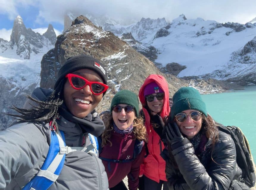 Un voyage en groupe WeRoad de quatre femmes souriantes prenant un selfie devant un lac turquoise et des montagnes enneigées.