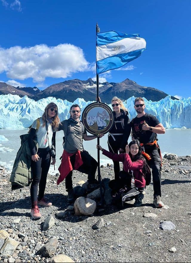 Un groupe WeRoad en voyage pose pour une photo avec un drapeau argentin et un panneau de parc devant un glacier immense.