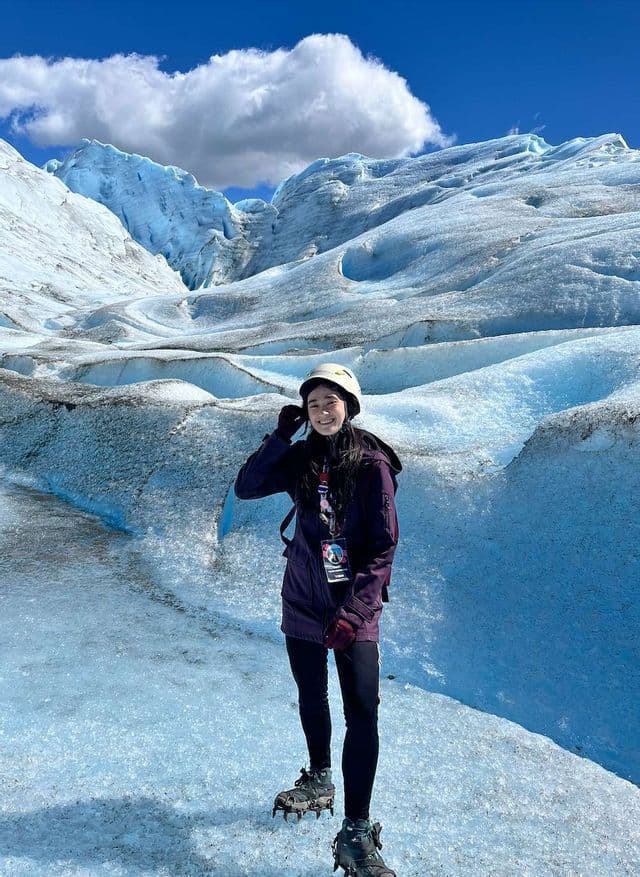 Une femme souriante, munie d'un casque et de crampons, se tient sur un vaste glacier de glace sous un ciel bleu clair.