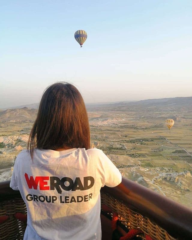Un Chef de Groupe WeRoad, vu de dos, admire depuis une montgolfière un vaste paysage avec d'autres ballons dans le ciel.