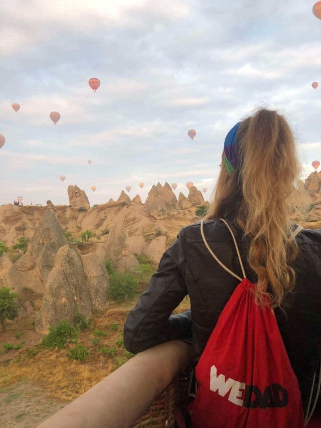 Una donna con una borsa WeRoad in viaggio osserva da una mongolfiera altre che fluttuano sopra una valle rocciosa.