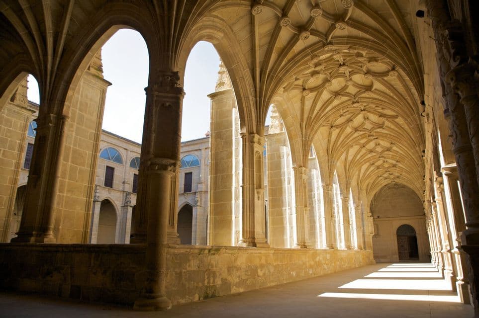 Un couloir de pierre ensoleillé d'un bâtiment historique, avec des arches gothiques et un plafond voûté orné, donnant sur une cour centrale.
