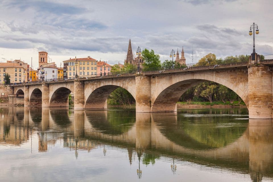 Un puente de piedra con múltiples arcos cruza un río en calma, con el horizonte de una ciudad histórica reflejado en el agua bajo un cielo nublado.