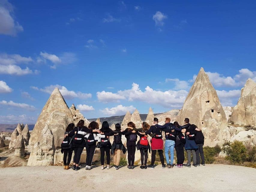 Rückansicht einer WeRoad-Gruppenreise, bei der sich die Teilnehmer umarmen und konische Felsformationen unter blauem Himmel bewundern.