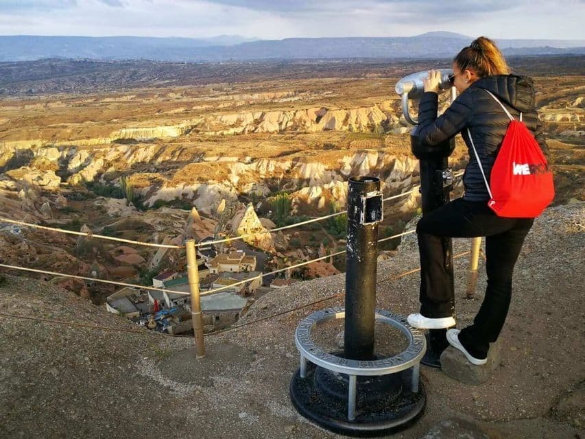 Una donna con uno zaino WeRoad rosso guarda attraverso un mirino, ammirando una vasta valle rocciosa da un punto panoramico elevato.