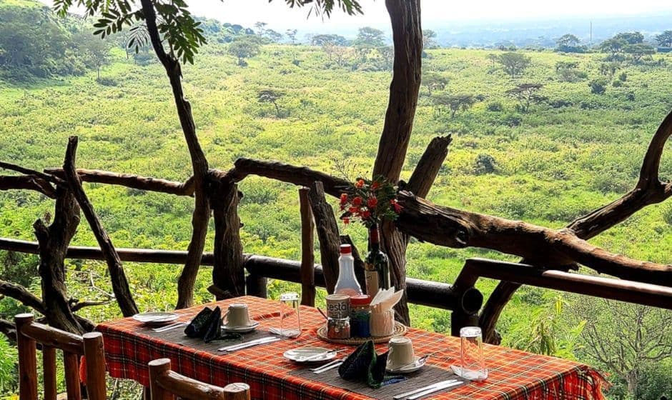 Un tavolo da pranzo con tovaglia a quadri apparecchiato per un pasto su un balcone rustico in legno con vista su una vasta savana verde.