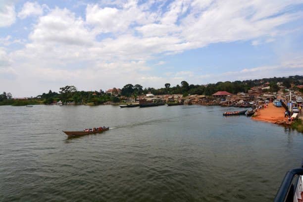 Una lunga barca di legno con diverse persone si muove sull'acqua verso un vivace villaggio sulla riva sotto un cielo nuvoloso.