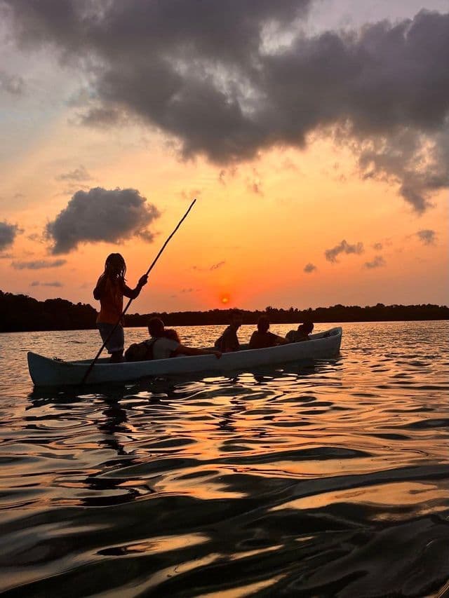Una persona rema una canoa che trasporta un viaggio di gruppo WeRoad sull'acqua, stagliata contro un tramonto nuvoloso e arancione.