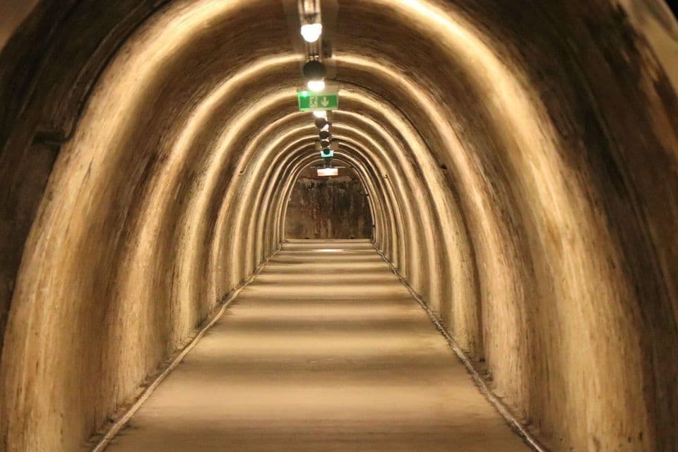 Un lungo tunnel sotterraneo ad arco con pavimento in cemento, illuminato da una fila di luci a soffitto che creano una prospettiva sfuggente.