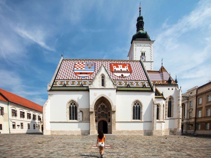 A woman walks across a cobblestone square towards a white church with a colorful, patterned tile roof under a blue sky.