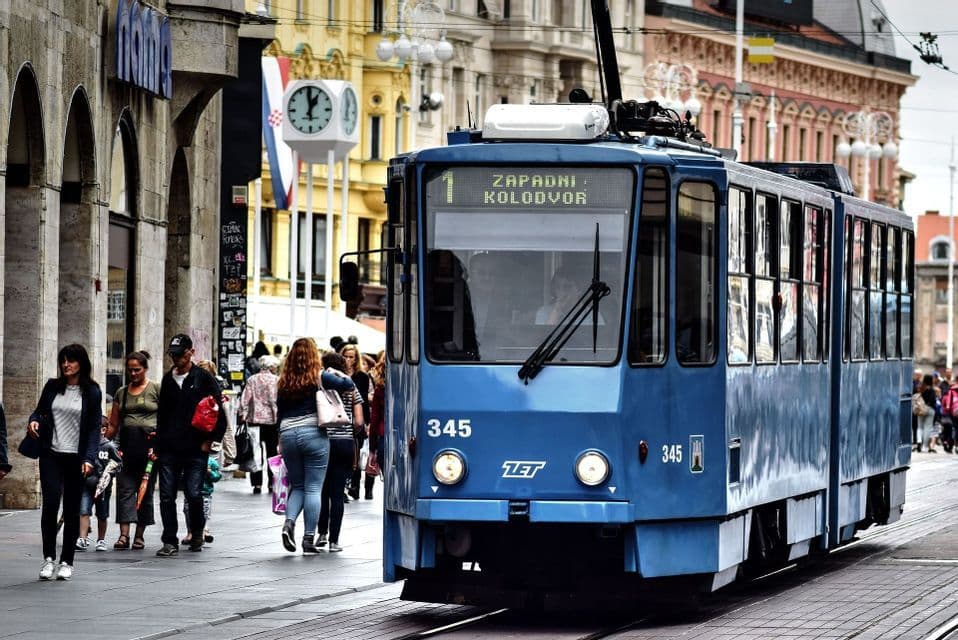 A blue tram travels on tracks down a busy city street as people walk along the sidewalk.