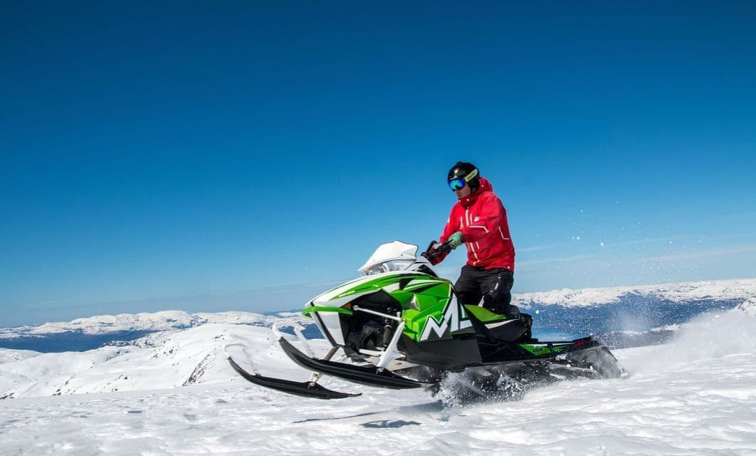 Una persona con chaqueta roja y casco conduce una moto de nieve verde por un paisaje montañoso nevado bajo un cielo azul claro.