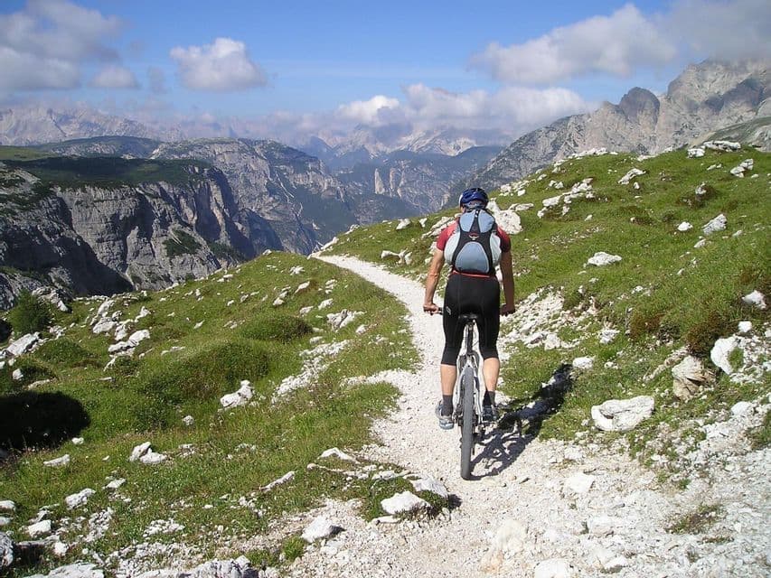 Ciclista de montaña en un sendero estrecho a lo largo de una ladera cubierta de hierba, con un vasto valle montañoso de fondo.