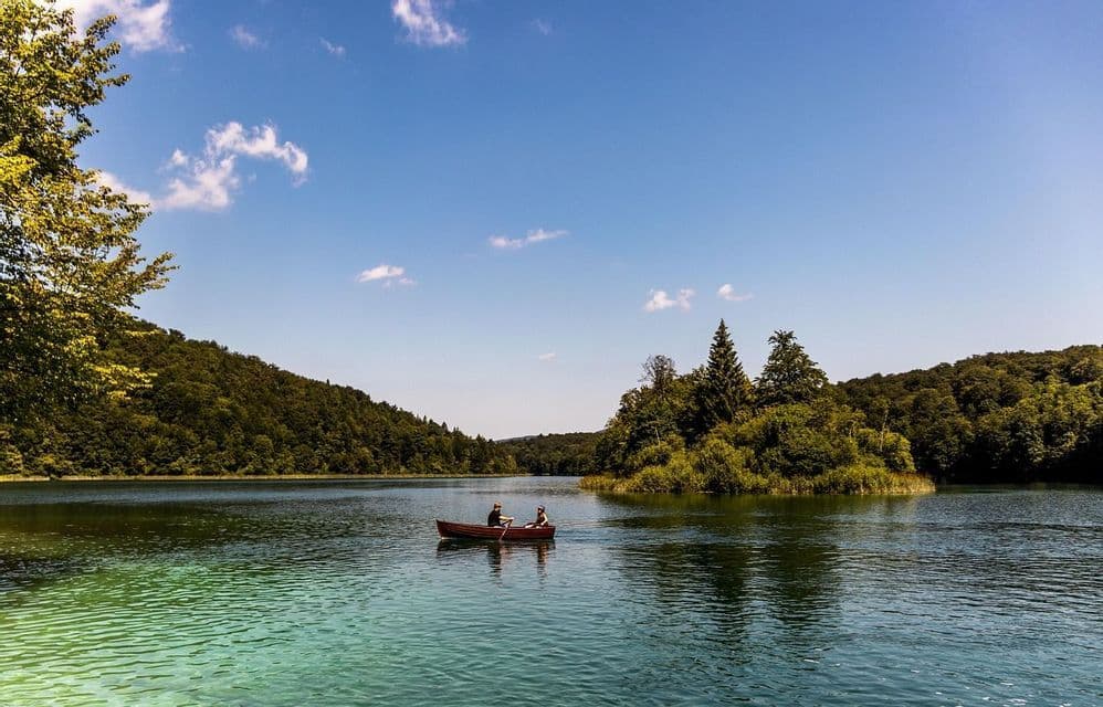 Due persone in un viaggio di gruppo WeRoad remano una barca di legno attraverso un lago calmo e turchese circondato da foreste verdi.