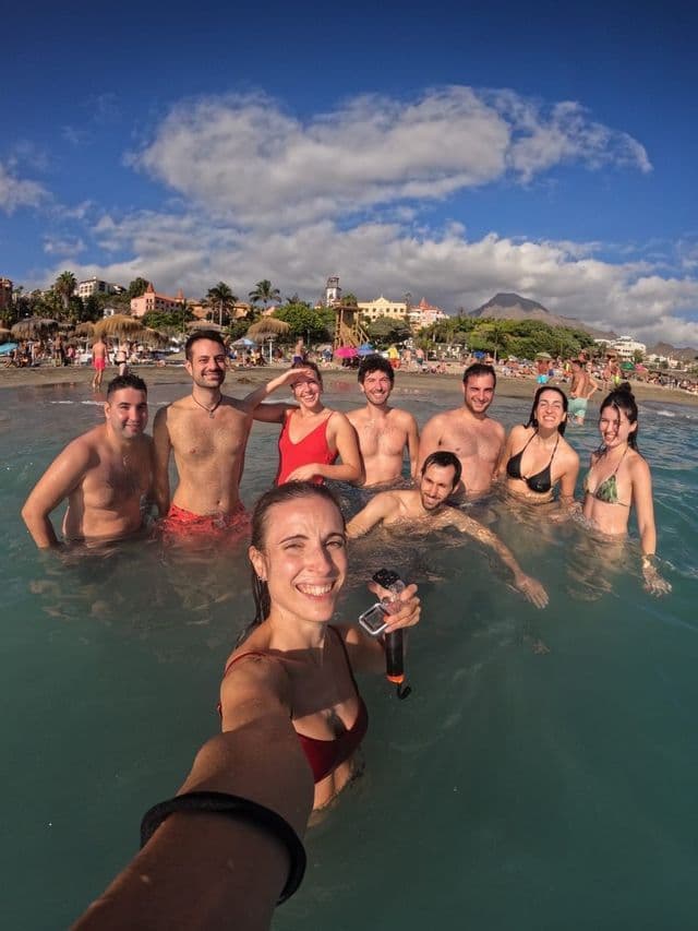 Eine Frau macht ein Selfie mit ihrer WeRoad-Gruppe, während sie an einem überfüllten Strand unter teilweise bewölktem Himmel im Meer schwimmt.
