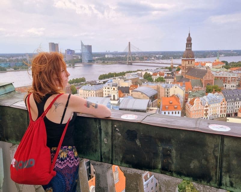 Eine Frau mit roten Haaren und rotem Rucksack lehnt an einer Brüstung und blickt über eine Stadtsilhouette mit Fluss und Kirchturm.