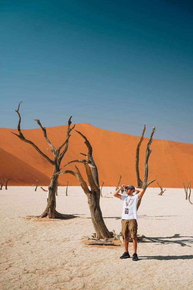 Ein Mann steht inmitten toter Bäume auf einer weißen Lehmpfanne vor einer massiven orangefarbenen Sanddüne unter klarem Himmel.