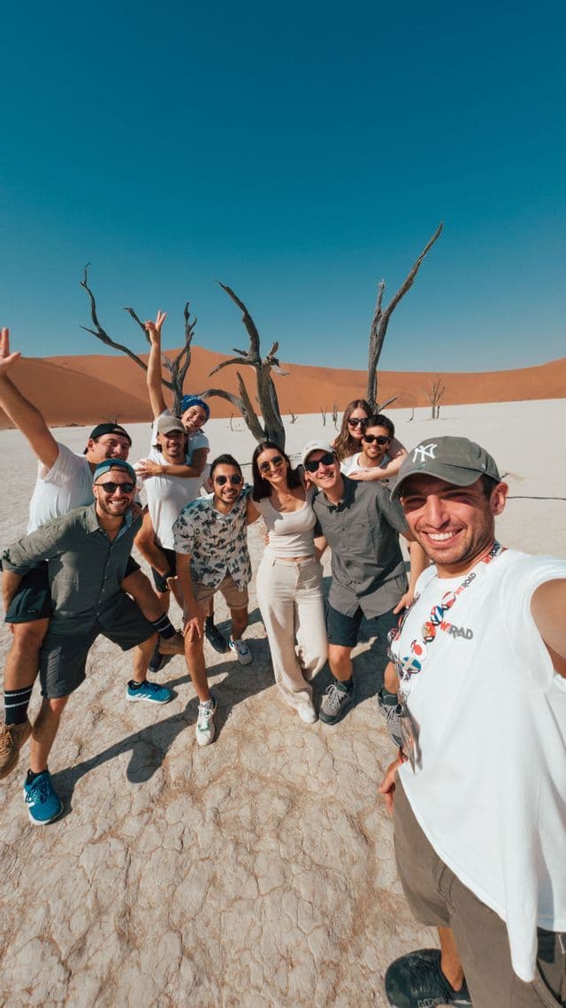 Un gruppo WeRoad si scatta un selfie sorridente in un deserto arido, con alberi scheletrici e dune di sabbia arancioni sullo sfondo.