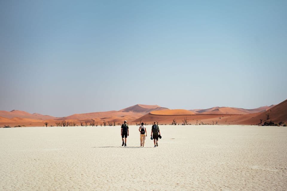 Drei Personen einer WeRoad Gruppenreise gehen unter einem klaren blauen Himmel über eine weiße Salzebene auf große orangefarbene Sanddünen zu.