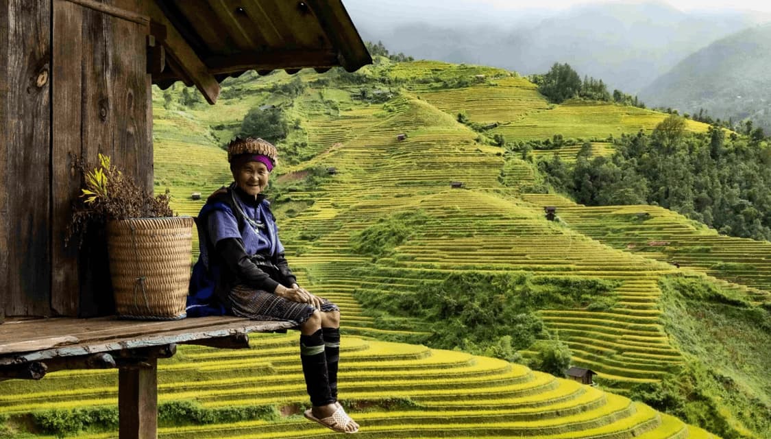 An elderly woman in traditional clothing sits smiling on a wooden porch overlooking vast terraced rice paddies.