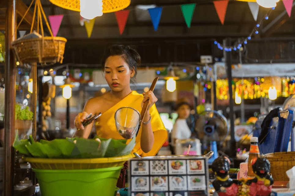 A woman in a traditional yellow dress prepares food with a ladle at a bustling market stall decorated with colorful bunting.