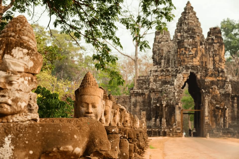 A row of ancient stone statues with serene faces lines a path leading towards a large temple gate surrounded by trees.