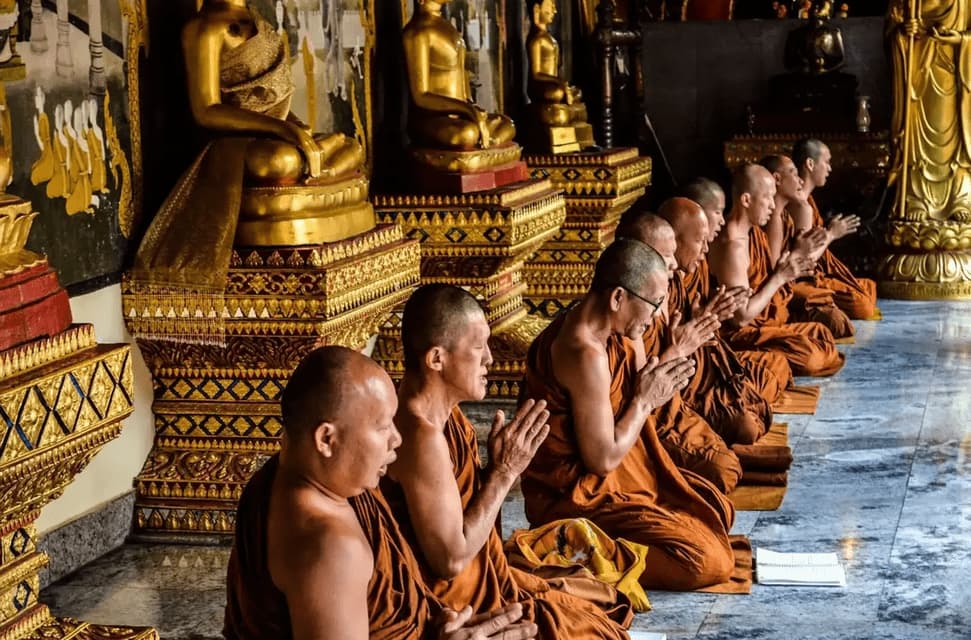 A line of Buddhist monks in orange robes kneeling to pray inside a temple in front of golden Buddha statues.