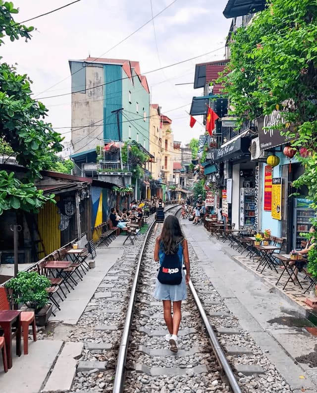 A person wearing a WeRoad backpack walks on a railway track through a narrow street lined with cafes and buildings.
