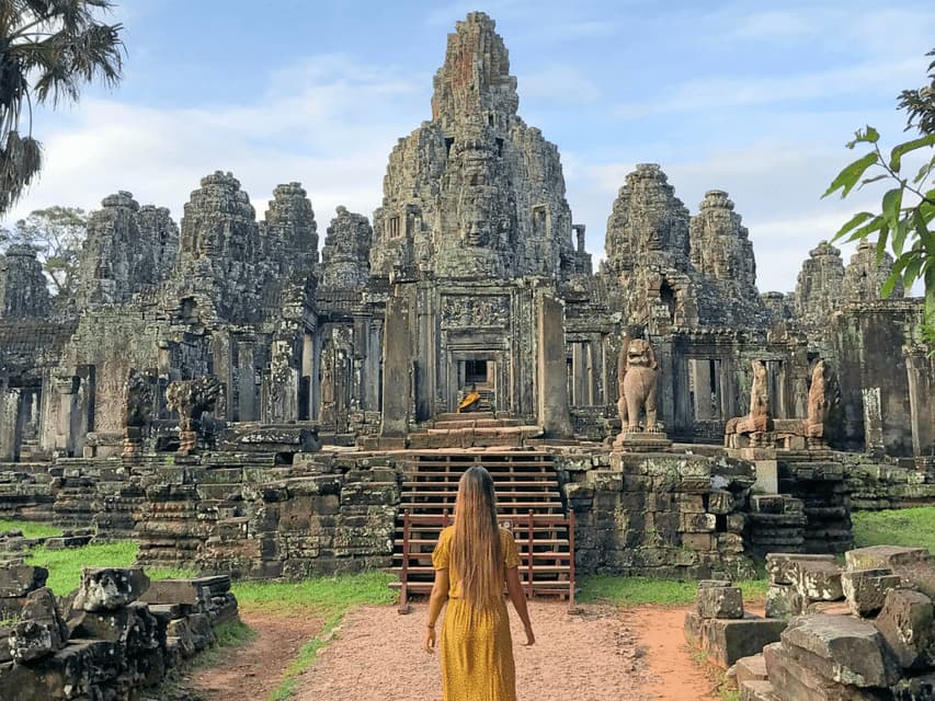 A woman with long hair in a yellow dress walks on a path towards the entrance of a large, ancient stone temple with carved towers.