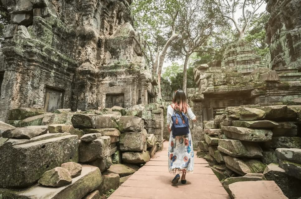 A woman with a blue backpack walks on a wooden pathway through ancient, moss-covered stone temple ruins.