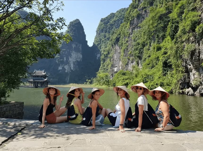 A WeRoad group trip of women wearing conical hats sits by a lake, with a pagoda and lush green mountains in the background.
