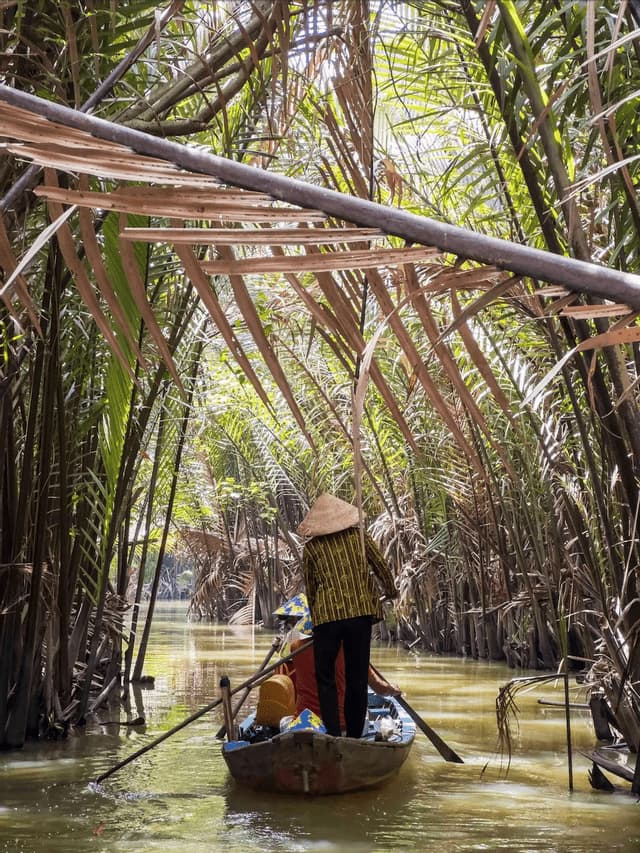 A person in a conical hat rows a small boat with a passenger down a narrow river flanked by dense palm trees.