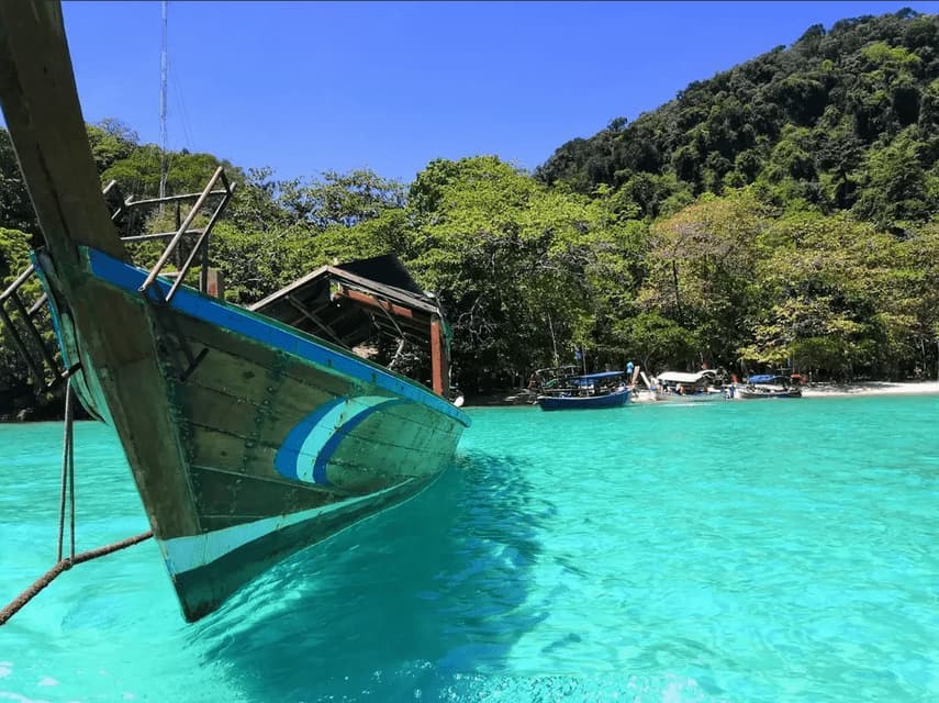 The bow of a wooden boat moored in vibrant turquoise water, with a tropical, tree-covered island in the background.