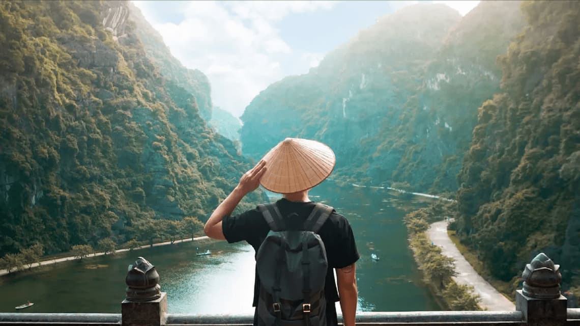A person from behind wearing a conical hat and backpack looks out over a river valley between steep, green mountains.