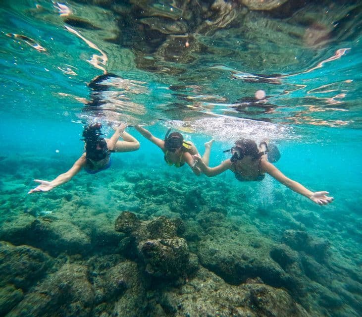 Three people from a WeRoad group trip snorkeling together, holding hands in clear turquoise water above a rocky seabed.