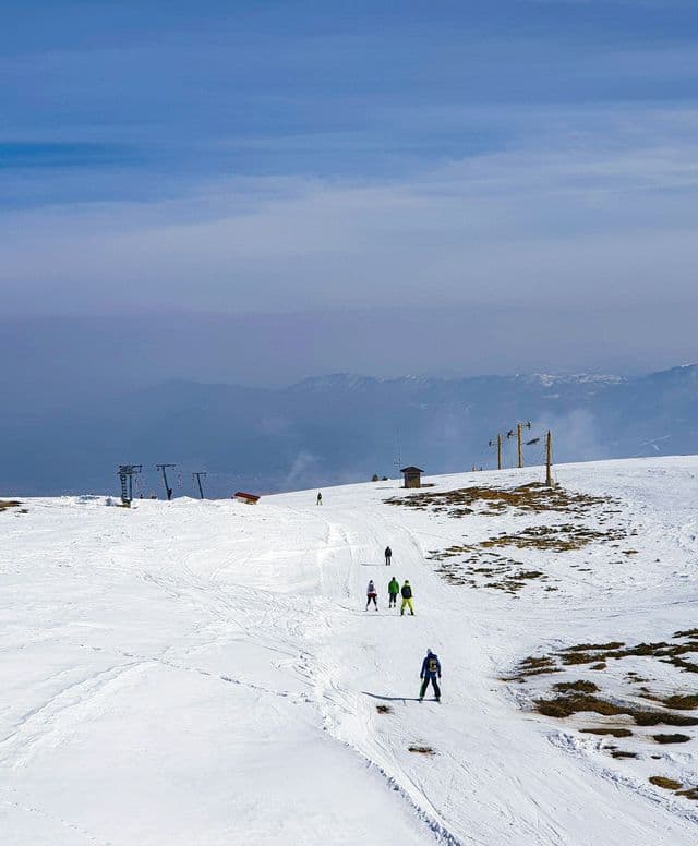Skifahrer und Snowboarder auf einem breiten, schneebedeckten Hang an einem Berg unter dunstigem Himmel.