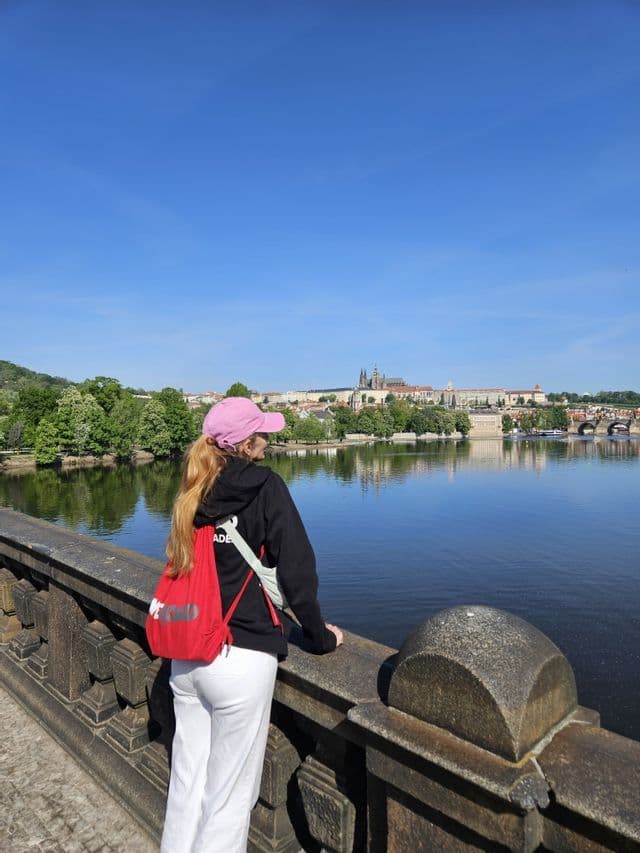 Une femme portant une casquette rose et un sac à dos rouge se tient sur un pont de pierre, observant une rivière en direction d'un paysage urbain.