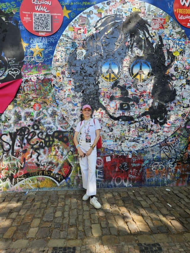 Une femme vêtue de blanc et coiffée d'une casquette rose sourit devant le mur Lennon coloré, couvert de graffitis et d'autocollants.