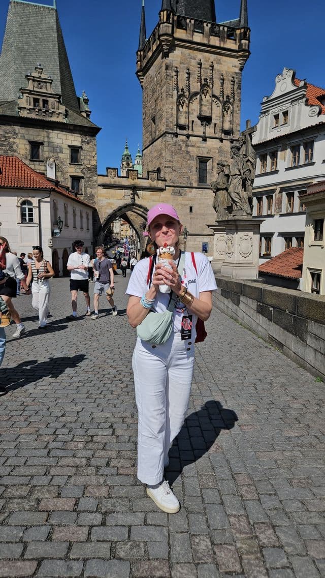 Une femme en voyage WeRoad tient un cornet de dessert sur un pont pavé ensoleillé, avec des tours historiques en arrière-plan.